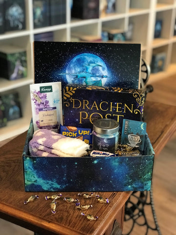 Das Produktbild zeigt vorn eine geöffnete Drachenpost in blauem Schuppen-Design. Man sieht im Deckel der Box das Verlagslogo - einen Drachen, der vor dem Mond fliegt - und in der Box die zum Thema Drachentraum passenden Goodies - Kuschelsocken, Badezusatz, Gute-Nacht-Tee und andere Kleinigkeiten - sowie die Kerze "Fly me to the Moon" und ein in ein lilafarbenes Drachenpost-Papier eingepacktes Buch. Im Hintergrund sieht man die Bücherregale des Verlagsladens.